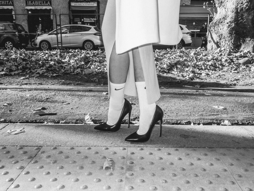 Black and white photograph focusing on the lower half of an individual. The person is wearing white ribbed socks paired with sleek black high heels. The scene captures the contrast between urban decay and personal style, as the neatly dressed legs juxtapose against the background filled with fallen leaves, a rugged road, and a parked car. The tactile pavement also adds an interesting foreground element, giving a sense of place and suggesting a crosswalk or pedestrian area.
