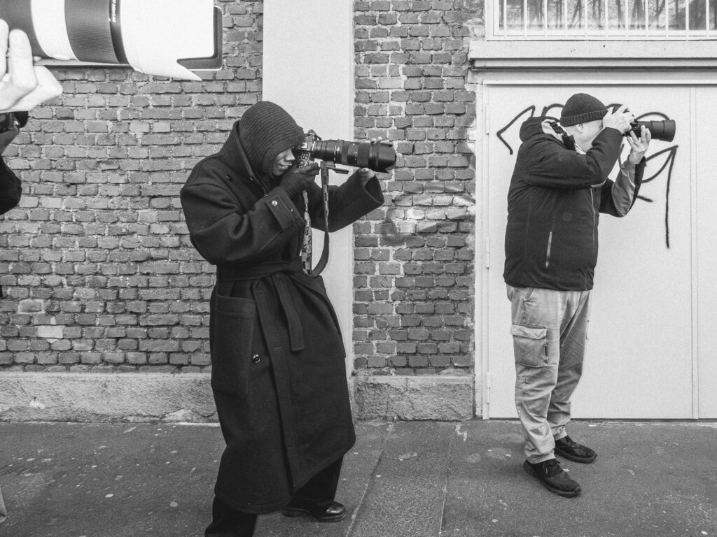 Monochromatic photograph capturing a candid moment of two photographers in action. One individual, cloaked in a dark hooded coat, is seen taking a photograph with a smile peeking from beneath the hood. The other person, dressed in casual attire with a beanie, focuses intently through his lens. A contrasting background of an old brick wall and a graffitied garage door encapsulates them. The shot subtly reveals the interplay of capturing and being captured, as another lens is seen in the foreground, suggesting the presence of a third photographer.