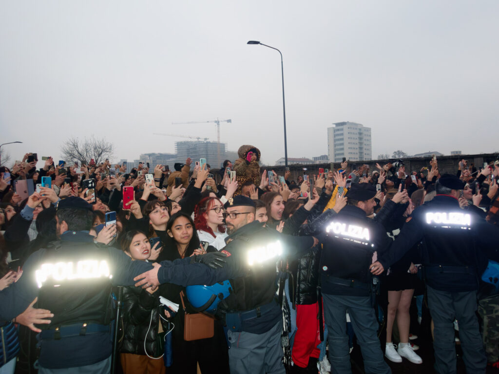 A crowd of people is captured holding up smartphones to take pictures or record an event. Four police officers with "POLIZIA" on their jackets are seen facing the crowd, seemingly there to maintain order. The crowd is diverse, with various expressions of excitement. The backdrop features a cityscape with construction cranes, indicating an urban setting at dusk or on a cloudy day.