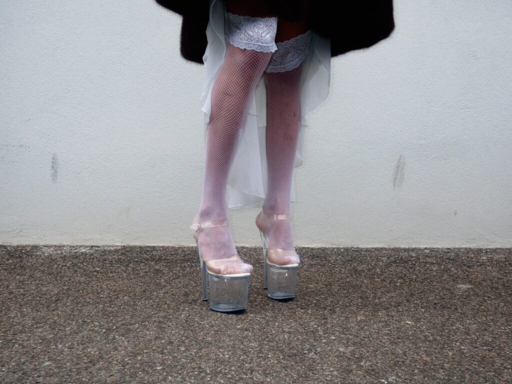 A close-up of a person's legs wearing white fishnet stockings, lace garters, and clear platform heels.