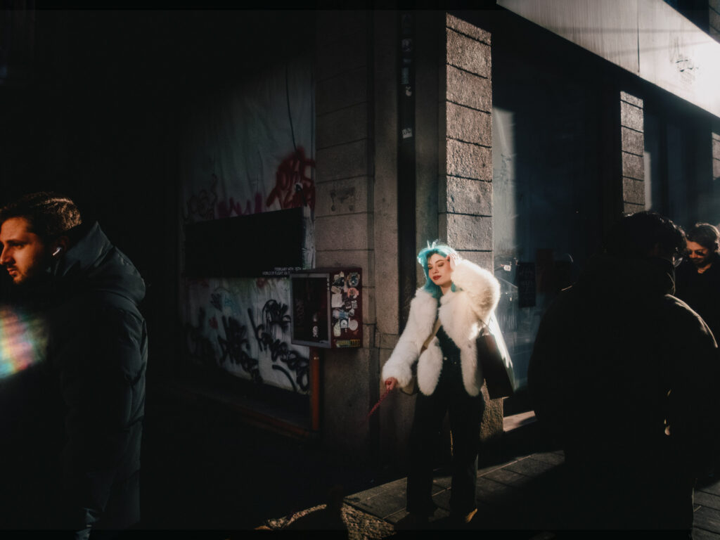 A street scene with dramatic lighting: a woman with blue hair and a white fur coat is illuminated by sunlight, standing against a graffiti-covered wall while holding a red leash. Shadows and dark-clad pedestrians frame the composition.