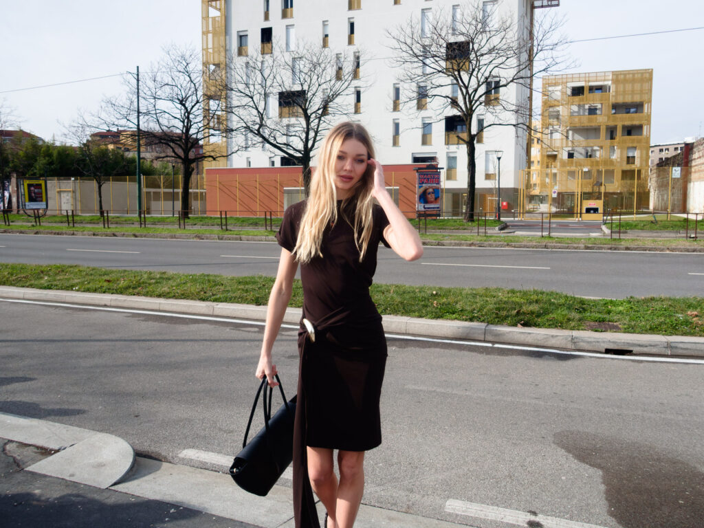 A woman in a black dress stands on a sidewalk with one hand adjusting her hair and the other holding a black bag. Behind her, a cityscape with modern buildings featuring geometric designs matching scene colors.