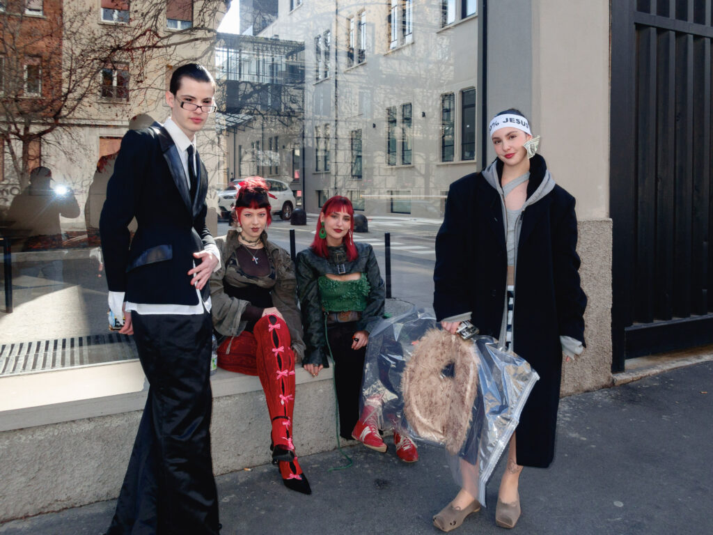 A group of four individuals posing on a street corner with a fashionable and eclectic style. On the left, a person in a tailored black suit with satin accents and glasses stands confidently. Next to them, two individuals with vibrant red hair sit on a low ledge, wearing bold outfits with intricate details like corset-style tops and bright red leggings. On the right, a person in an oversized black coat, hoodie, and headband reading '100% JESUS' holds a furry item in a clear plastic bag. The urban background features modern buildings with reflective windows.