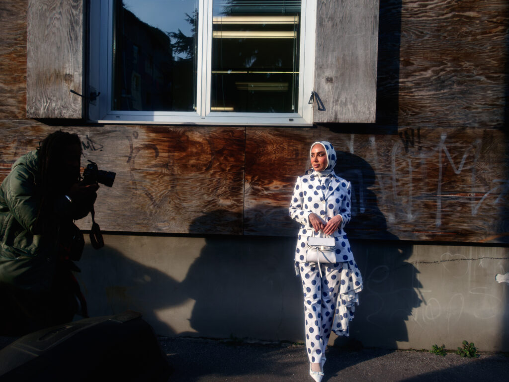 A fashionable attendee dressed in a white polka-dot ensemble with a matching headscarf poses against a weathered wooden wall during Milan Fashion Week. She carries a small white handbag and stands in the warm afternoon light, while a photographer crouches to capture her portrait, emphasizing the interplay of shadows and textures.