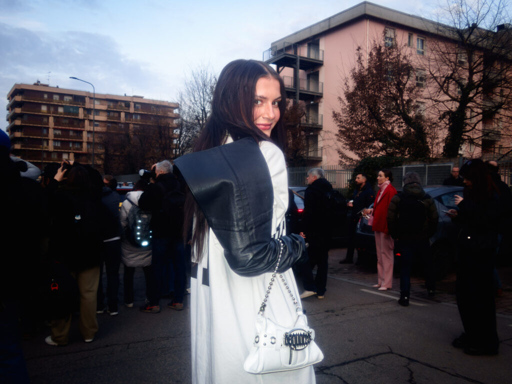 A stylish attendee with long dark hair glances back over her shoulder, wearing an oversized white top with a sculptural black leather sleeve. She carries a white handbag featuring metallic details, standing amidst a street crowd during Milan Fashion Week. In the background, pink and beige apartment buildings frame the scene.