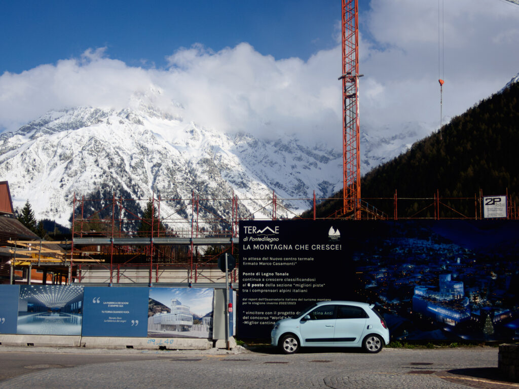 This image captures a striking contrast between nature and development. In the foreground, there's a construction site with scaffolding and a large crane. Promotional banners around the site announce a new thermal center, with renderings of the future building. A small light blue car is parked in front, accentuating the scale of the construction. Behind this scene of progress, the majestic, snow-capped mountains stand tall, underscoring the juxtaposition of human activity against the backdrop of the natural world.