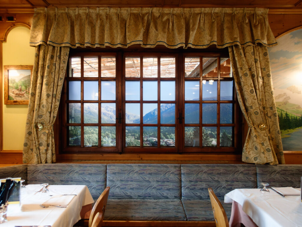 Inside a mountain-themed restaurant, a large window with patterned curtains reveals not the outdoors, but a wall-sized poster simulating a mountain view. The dining area is inviting, with white tablecloths on the tables and benches along the wall, all contributing to a charming and rustic atmosphere that evokes the essence of a cabin retreat.