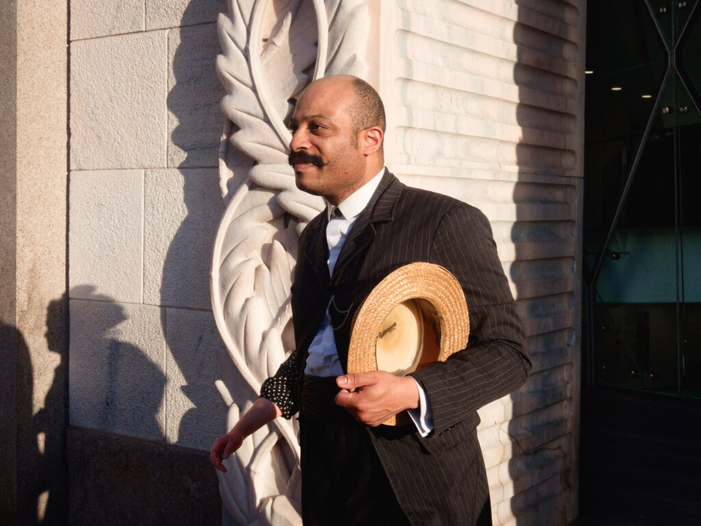 A man in a pinstripe suit holds a straw hat as he stands in front of an intricately carved stone wall. The scene is well-lit with natural sunlight, casting distinct shadows on the wall. The man's expression is calm and he appears to be mid-step. A woman's arm, visible in the background, suggests she is walking with him.