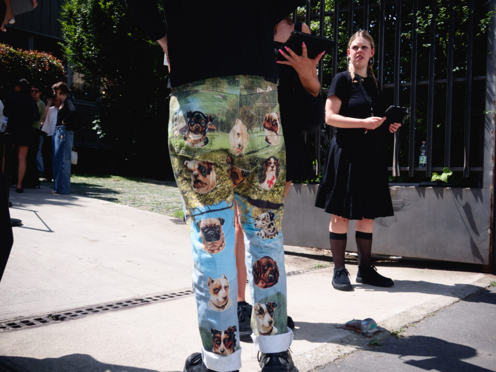 A street photography scene depicting a man wearing pants with vibrant dog portraits printed on them, standing in the foreground. He is accompanied by a woman dressed in a black skirt and top, holding a tablet, looking to her side with an expressive face.