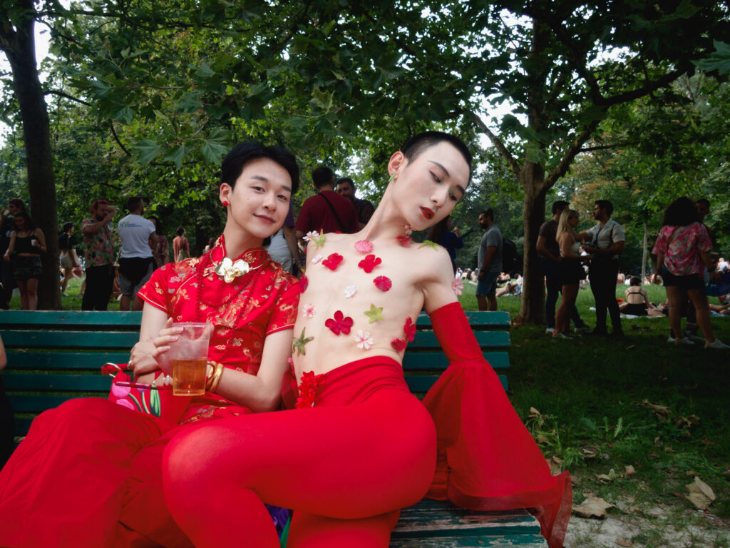 Two individuals dressed in striking red outfits sit on a park bench during Milano Pride 2024. The person on the right poses confidently, adorned with floral decorations on their upper body. The background features other attendees enjoying the event amidst the greenery of the park.