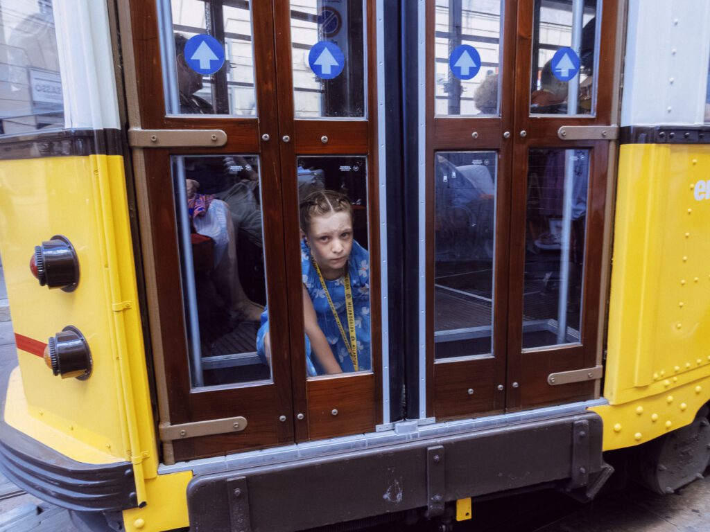 Image capturing a tram. Through the window, a young girl with braids is seen looking directly into the camera with a thoughtful expression. The tram doors display directional arrows.
