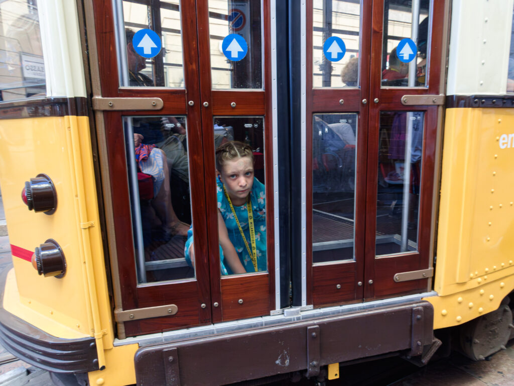 Black and white image capturing a tram. Through the window, a young girl with braids is seen looking directly into the camera with a thoughtful expression. The tram doors display directional arrows.
