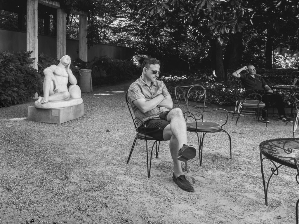 Black and white image of an outdoor setting with a stone sculpture of a reclining figure on a pedestal to the left. A man in sunglasses and casual attire sits contemplatively on an iron chair, while another individual relaxes on a chair in the background amidst lush vegetation.