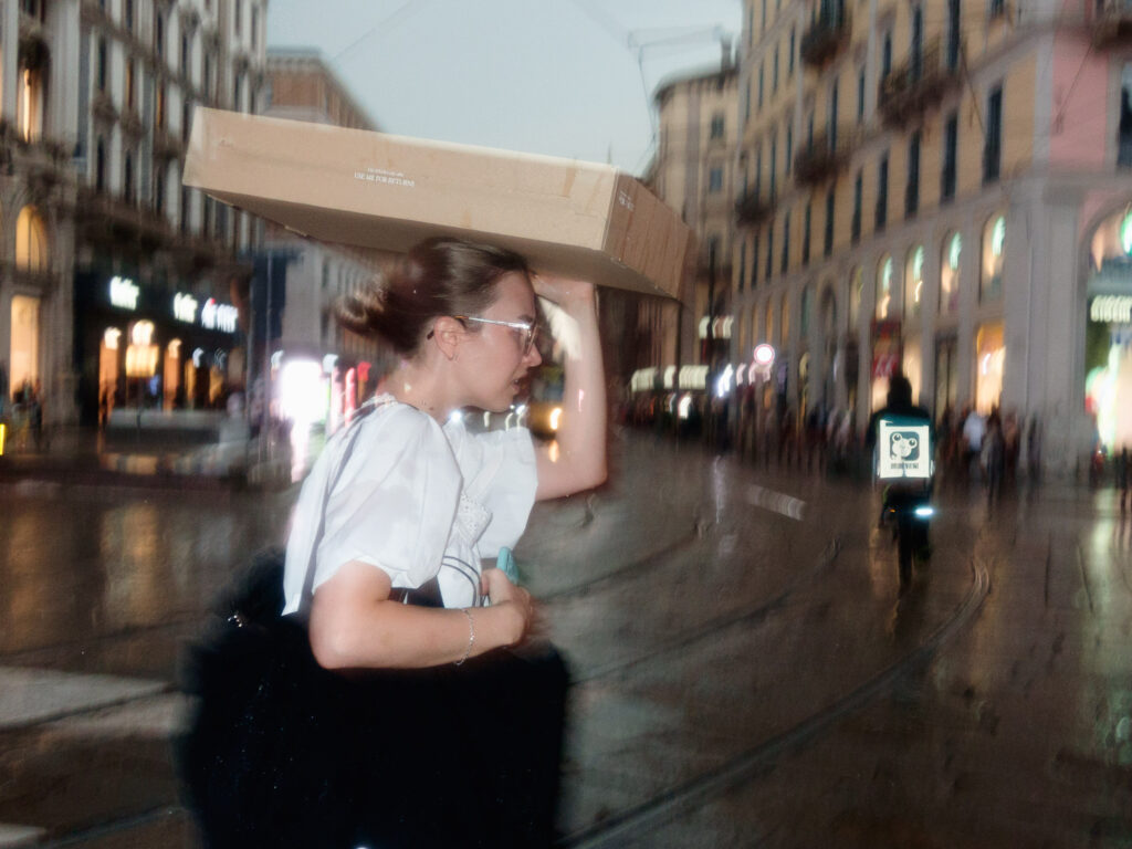 A woman carrying a cardboard box on her head walks through a busy, rain-soaked urban street at dusk, with blurred lights and buildings in the background.