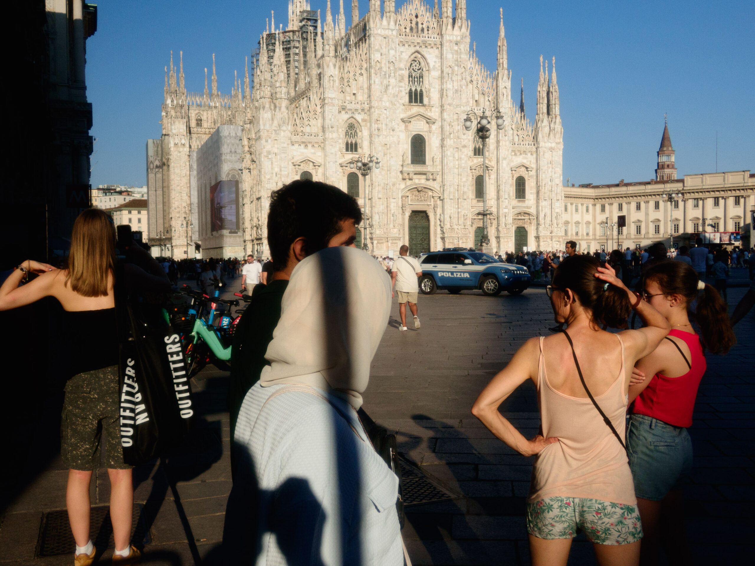 A bustling scene in front of the Milan Cathedral (Duomo di Milano) in the late afternoon sun. The image features various people, including a woman in a black top with a large "Outfitters" shopping bag, a person with a white headscarf, and two women in summer attire standing with their backs to the camera. A police car is parked in the background, and the cathedral's stunning gothic architecture is illuminated by the sunlight.
