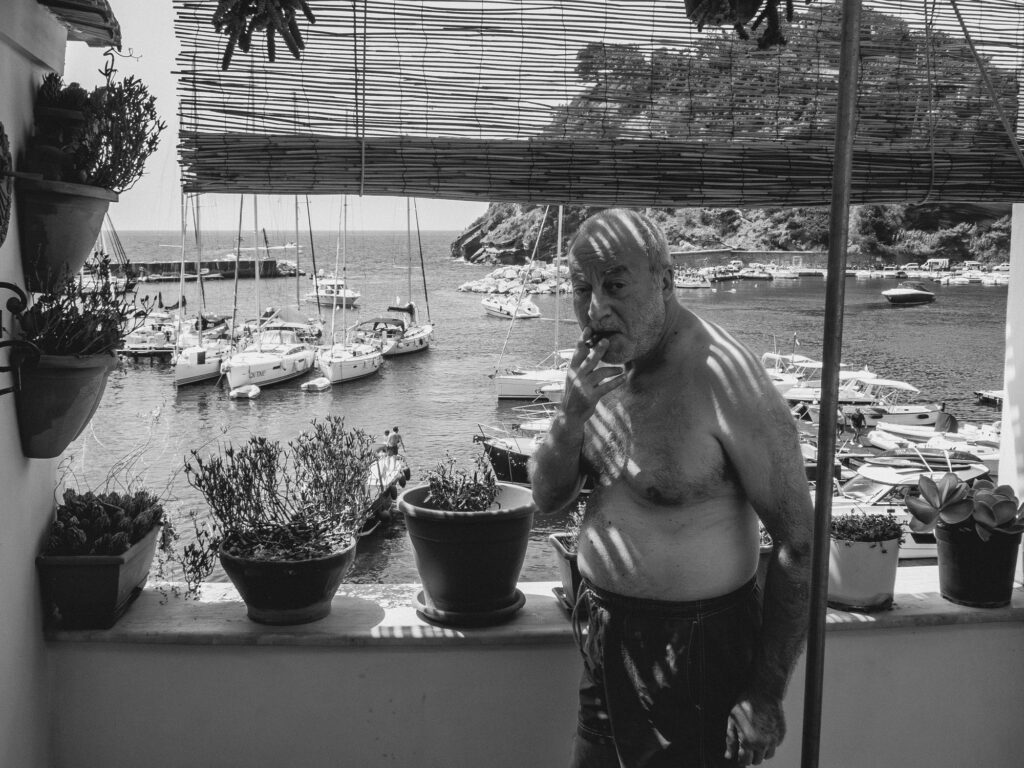 Photograph in black and white showcasing a seaside scene. In the foreground, an elderly man, bare-chested and in shorts, smokes a cigarette. His gaze is off-camera, creating a contemplative atmosphere. Behind him, through bamboo blinds, there is a picturesque view of a harbor filled with boats. Potted plants dot the balcony, adding to the Mediterranean ambiance.