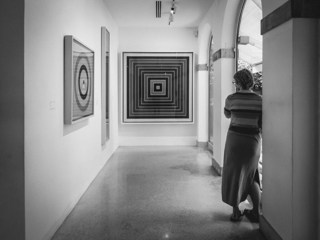 Black and white photograph depicting an art gallery setting. A woman in a striped top and long skirt attentively observes a geometric artwork on the wall. Other abstract artworks, characterized by concentric circles and squares, are visible in the softly lit space.