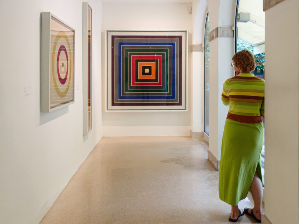 Black and white photograph depicting an art gallery setting. A woman in a striped top and long skirt attentively observes a geometric artwork on the wall. Other abstract artworks, characterized by concentric circles and squares, are visible in the softly lit space.