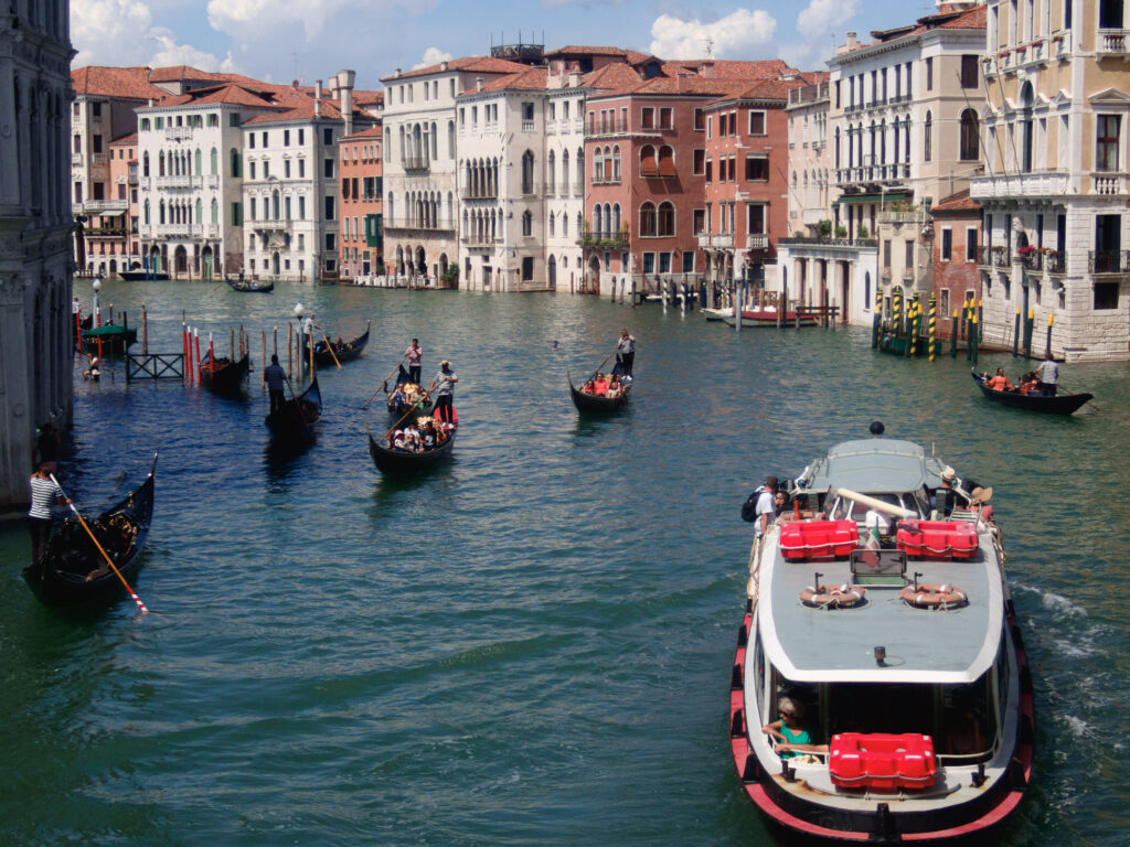 A vibrant scene on the Rialto Bridge in Venice. Multiple gondolas with gondoliers in traditional attire navigate the waterway, while a vaporetto, or water bus, occupies the foreground with passengers on board. Historic Venetian buildings line the banks, their facades reflecting the sunlight. The clear blue sky suggests a pleasant day, perfect for sightseeing in this iconic Italian city.