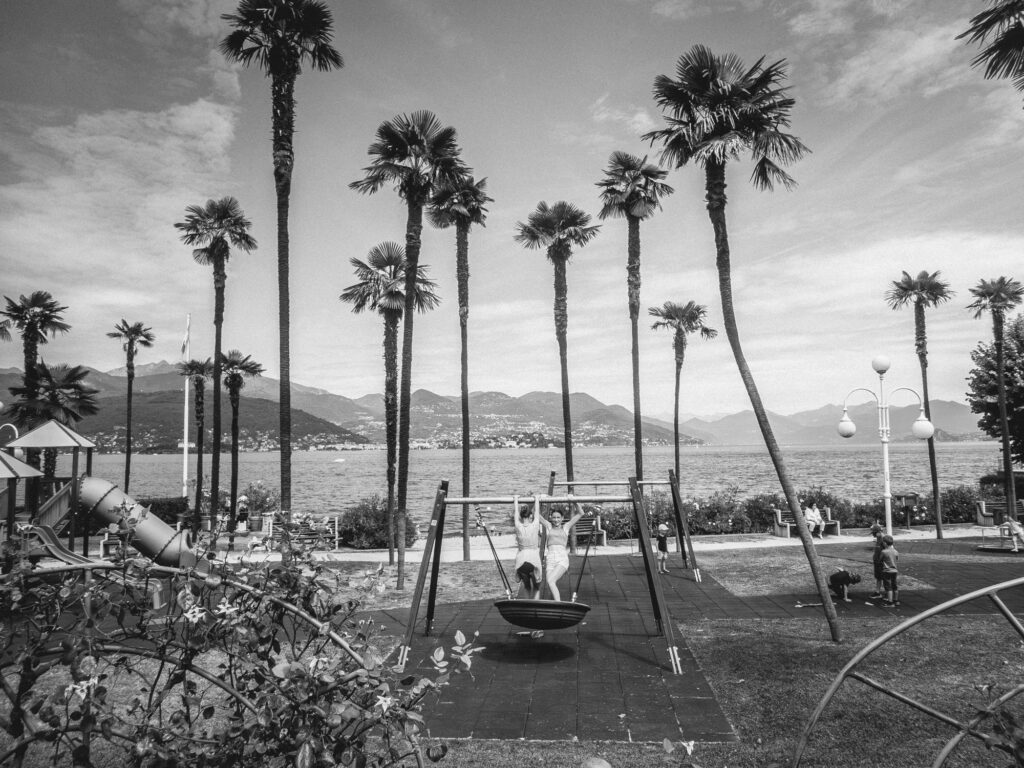 Black and white image of a coastal playground with tall palm trees lining the backdrop. In the foreground, children play on swings and a slide, while the distant mountains and calm waters of a lake or sea stretch out under a cloudy sky.
