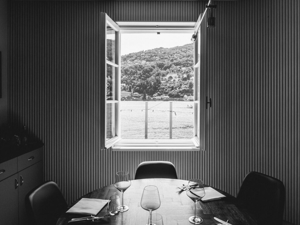 A black and white photograph captures a serene moment inside a dining area. The room features vertically striped walls that add depth and texture to the setting. At the center, a table is meticulously set with wine glasses, cutlery, and a folder or menu, suggesting an imminent meal or event. What stands out most, however, is the large window offering a breathtaking view of a lakeside landscape. Beyond the pane, a calm lake with distant boats and lush hills or mountains in the backdrop can be observed. The natural light streaming in gently illuminates the room, creating a relaxing and inviting ambiance. The entire composition evokes feelings of peace, reflection, and anticipation of a special moment.