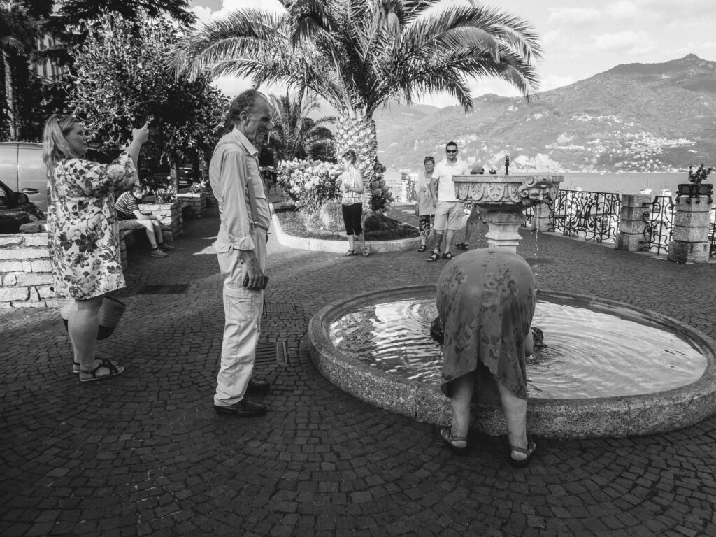 Black and white photograph of a moment at the Lake Como shoreline. A woman on the left is taking a photograph, while a distinguished-looking man in the center gazes at a woman who bends over a stone fountain, her face obscured by her own posture. In the background, two men stand by an ornate railing, overlooking the serene waters of Lake Como and the majestic mountains beyond. This scene captures a blend of daily life and the timeless beauty of the Italian lakeside setting.