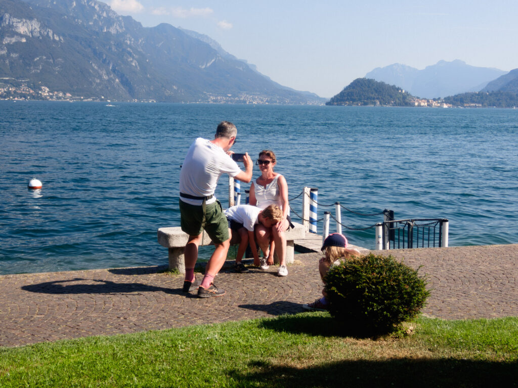 A serene lakeside scene featuring a family enjoying a sunny day. A man in shorts and a white t-shirt crouches with a smartphone, taking a photo of a woman sitting on a bench with her tired and exhausted young son resting on her legs. Nearby, a young girl wearing a hat sits on the ground. In the foreground, a small bush adds greenery, while the background showcases a stunning lake surrounded by majestic mountains under a clear blue sky.