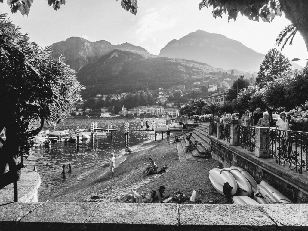 Black and white photo capturing a lively lakeside scene at Lake Como. People of various ages are enjoying the waterside, with some swimming in the lake, others sunbathing on the stone edge, and a few admiring the view from an ornate railing. Kayaks rest on the shore, waiting for their next journey. In the backdrop, majestic mountains tower over quaint buildings nestled on the hillside. Trees frame the view, offering shade and enhancing the scenic beauty. The atmosphere is one of relaxation and leisure, encapsulating the essence of a serene Italian lakeside town.