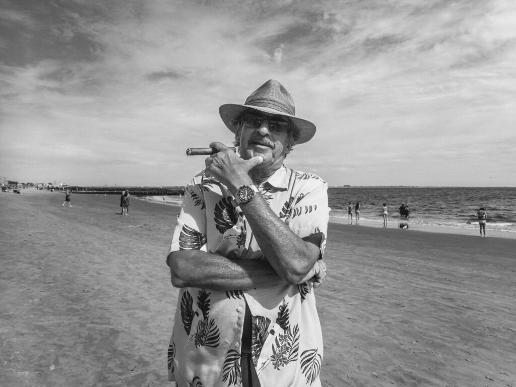 Black and white photograph of an older man standing on a beach. He wears a straw hat, sunglasses, and a tropical-patterned shirt. He is confidently smoking a cigar. The sandy beach stretches out behind him, dotted with a few distant figures and the shimmering sea. The sky above is spacious with a few clouds.