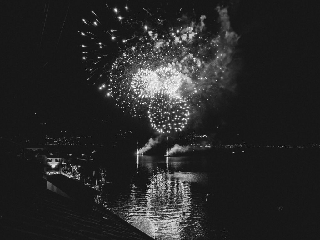 Black and white photo of a vibrant firework display emanating from two distant boats on Lake Como. The arrangement and shape of the fireworks bear a resemblance to a phallic symbol. The serene waters of the lake reflect the light and patterns of the fireworks, with the surrounding landscape dimly visible.