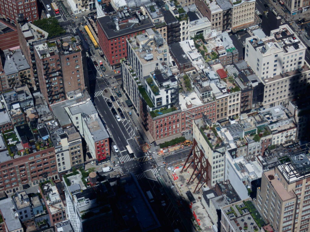 Aerial view of a dense urban cityscape, showcasing a patchwork of rooftops, streets with crosswalks, buildings of various heights, and scattered infrastructure elements.