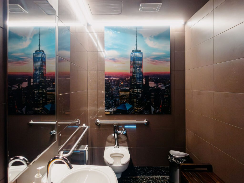 A modern bathroom featuring a large printed tile depicting a city skyline with a prominent skyscraper, alongside reflective surfaces, a sink, toilet, and grab bars.