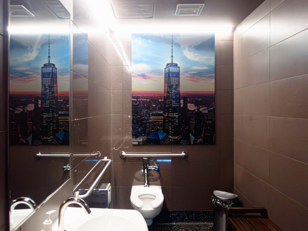A modern bathroom featuring a large printed tile depicting a city skyline with a prominent skyscraper, alongside reflective surfaces, a sink, toilet, and grab bars.