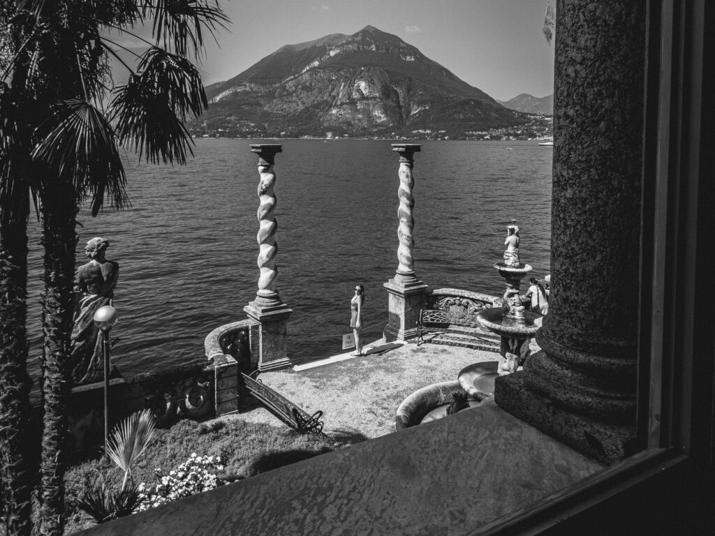 Photograph in black and white depicting a serene lakeside view taken from a window frame. Between two ornate, spiraled columns, stands a girl. Tall palm trees flank the left side, accompanied by stone sculptures. In the distance, mountains rise majestically above the calm lake waters, with small buildings visible along the shoreline.