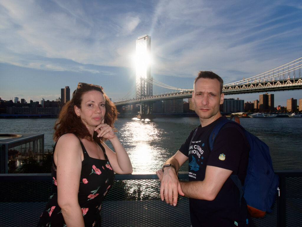 Photo of a woman with long hair, wearing a floral dress, and a man with a backpack standing by a waterfront, with a scenic view of a bridge and city skyline in the background.