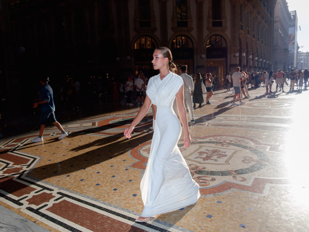 Street scene capturing a moment of contrast: a poised woman in an elegant, flowing dress stands prominently in the foreground, while in the backdrop, busy pedestrians walk across a decorative mosaic pavement amidst historic buildings bathed in sunlight.