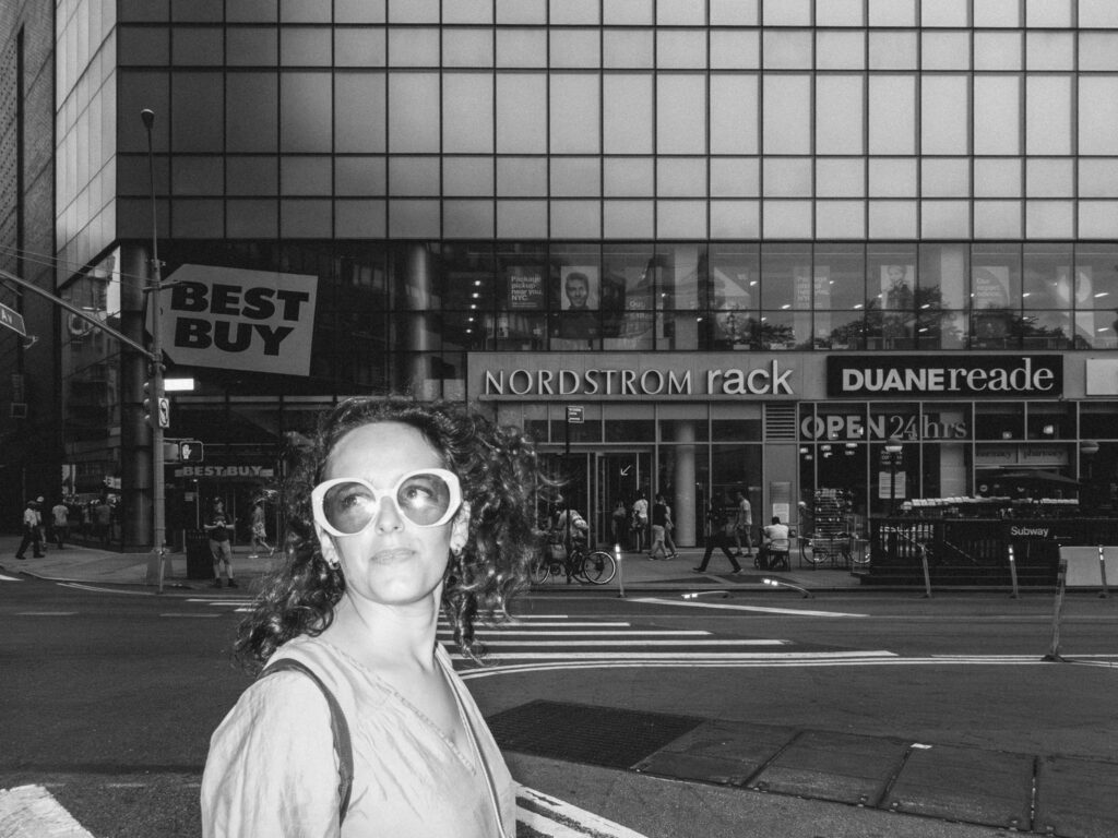 Black and white photograph of a woman with curly hair and large, round sunglasses in the foreground, standing against a bustling urban backdrop. Behind her, the facades of buildings display prominent signs for 'Best Buy', 'NORDSTROM rack', and 'DUANEreade'. Pedestrians cross the street, and city life unfolds under the reflection-laden windows of the adjacent building. The contrast between the woman's composed expression and the fast-paced city environment creates a compelling visual narrative.