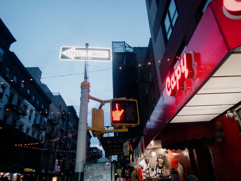 Vibrant urban street scene at dusk with a humorous twist, where the red hand of the pedestrian signal appears to be making a rude gesture, juxtaposed against the glowing 'ONE WAY' sign and neon-lit cafe signage.