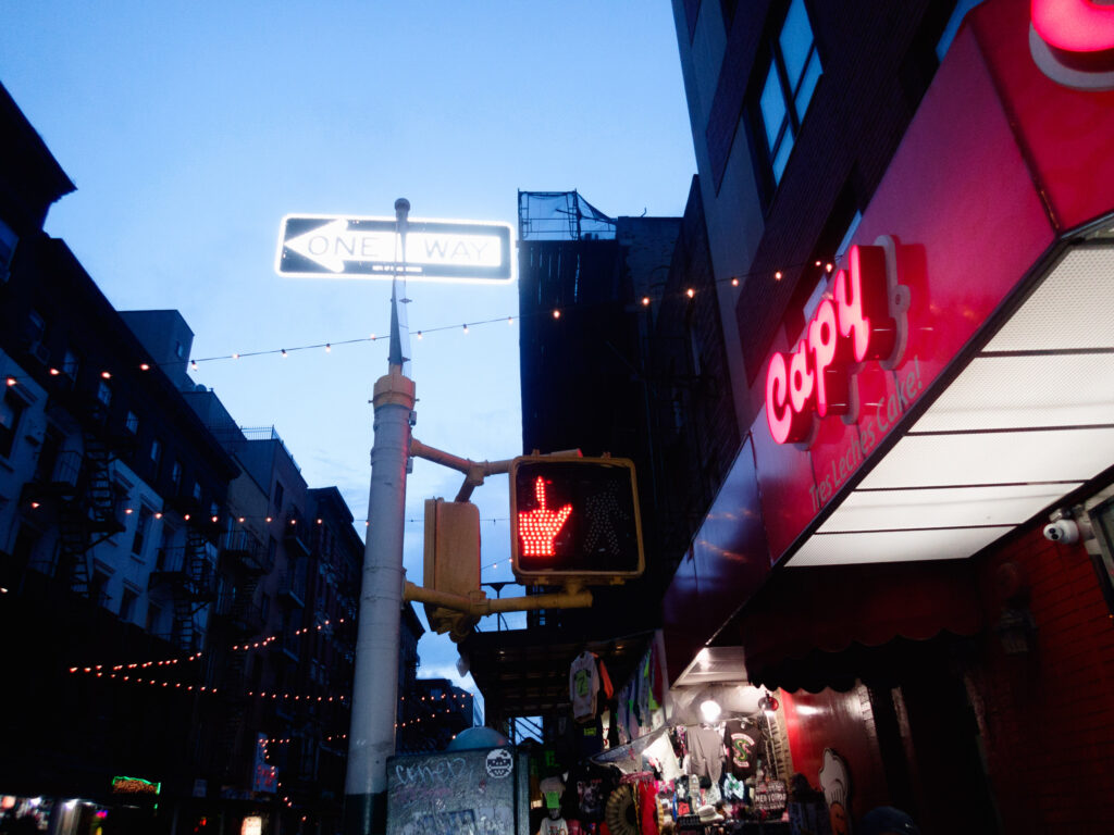Vibrant urban street scene at dusk with a humorous twist, where the red hand of the pedestrian signal appears to be making a rude gesture, juxtaposed against the glowing 'ONE WAY' sign and neon-lit cafe signage.
