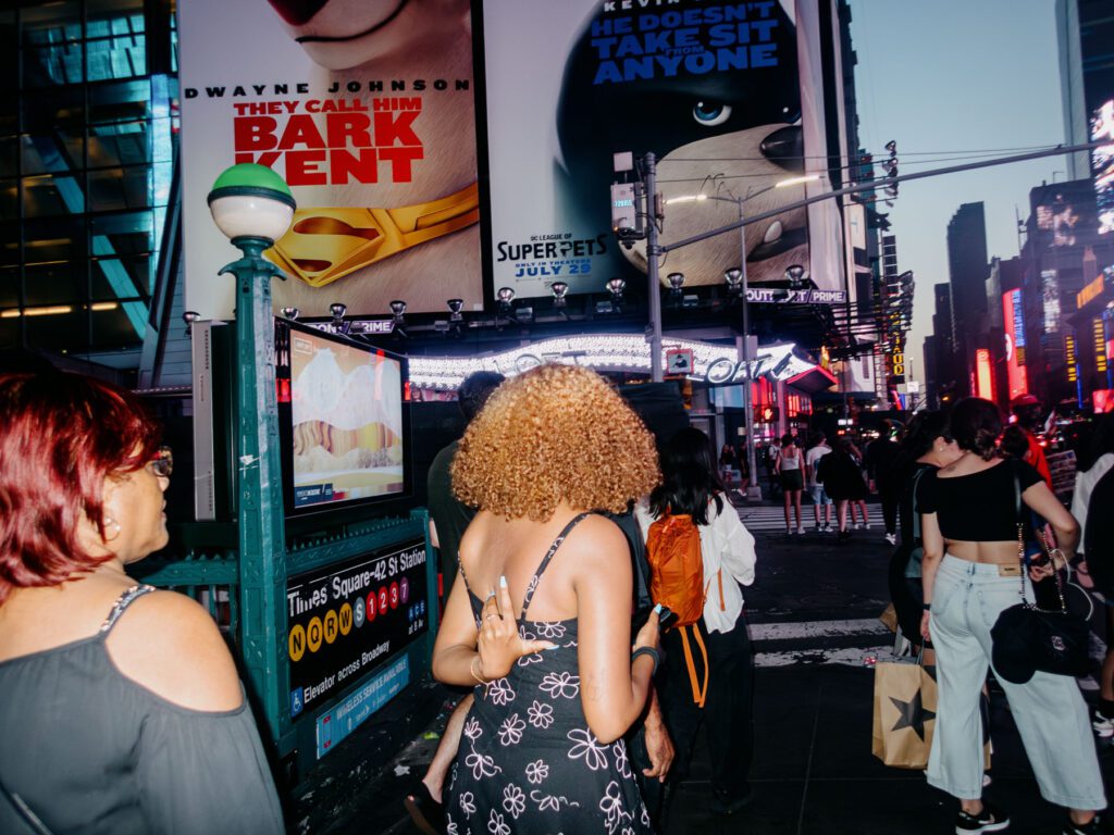 Photograph capturing a lively street scene at Times Square. People are milling about. The surroundings are illuminated by the bright lights of billboards and advertisements, including one featuring a parody of a superhero movie. The time of day appears to be evening, as indicated by the artificial lighting and active ambiance. The image conveys the hustle and bustle typical of this iconic New York City landmark.