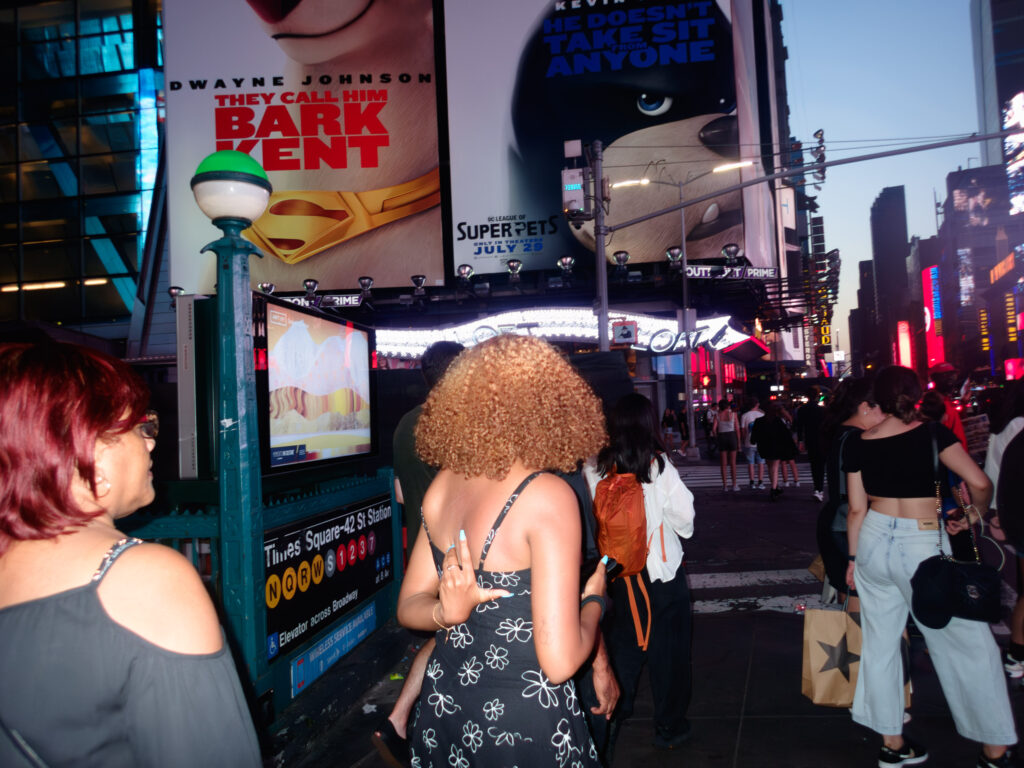 Photograph capturing a lively street scene at Times Square. People are milling about. The surroundings are illuminated by the bright lights of billboards and advertisements, including one featuring a parody of a superhero movie. The time of day appears to be evening, as indicated by the artificial lighting and active ambiance. The image conveys the hustle and bustle typical of this iconic New York City landmark.