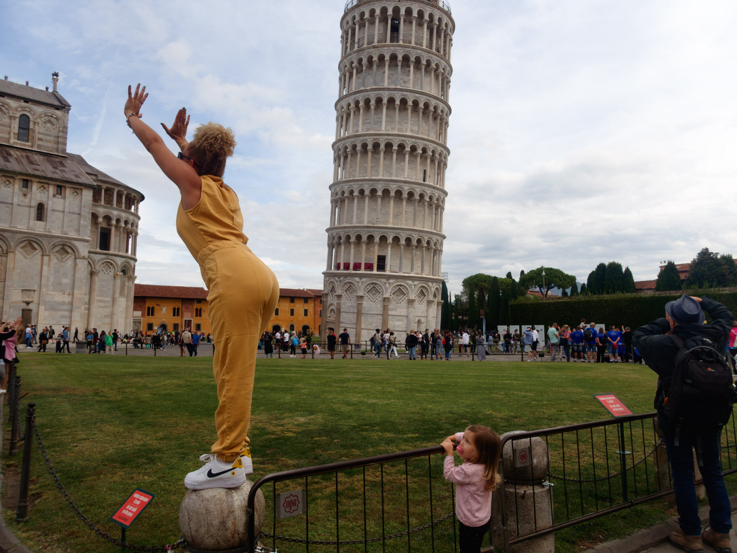 A striking photograph captures the essence of tourism and interaction at the iconic Leaning Tower of Pisa. A woman, prominently placed in the foreground, balances atop a pedestal attempting the classic pose of "holding up" the leaning tower, indulging in the playful optical illusion. In contrast, on the right, a young girl with eyes bright with curiosity holds up a pacifier, seemingly puzzled by the woman's pose. Against the backdrop, the Tower of Pisa stands majestic, with a crowd of tourists bustling about, each engrossed in their unique moment. The image is a captivating representation of the varied perspectives and interactions that converge at such an iconic spot.