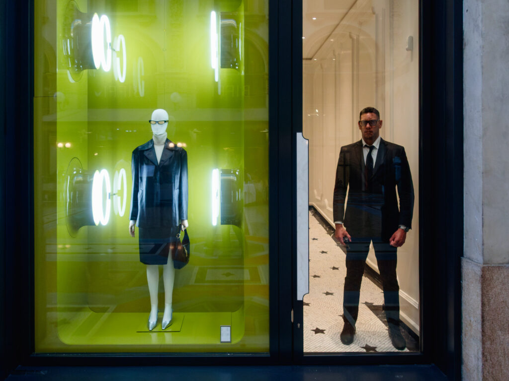 Color photograph of a store window at night. On the left, a female mannequin dressed in a chic leather jacket and skirt stands poised. On the right, a real male store employee in a sharp suit stands with a similar posture, blending in seamlessly as if he's a live counterpart to the mannequin. Neon lights reflect on the glass, creating a juxtaposition between the animate and inanimate.