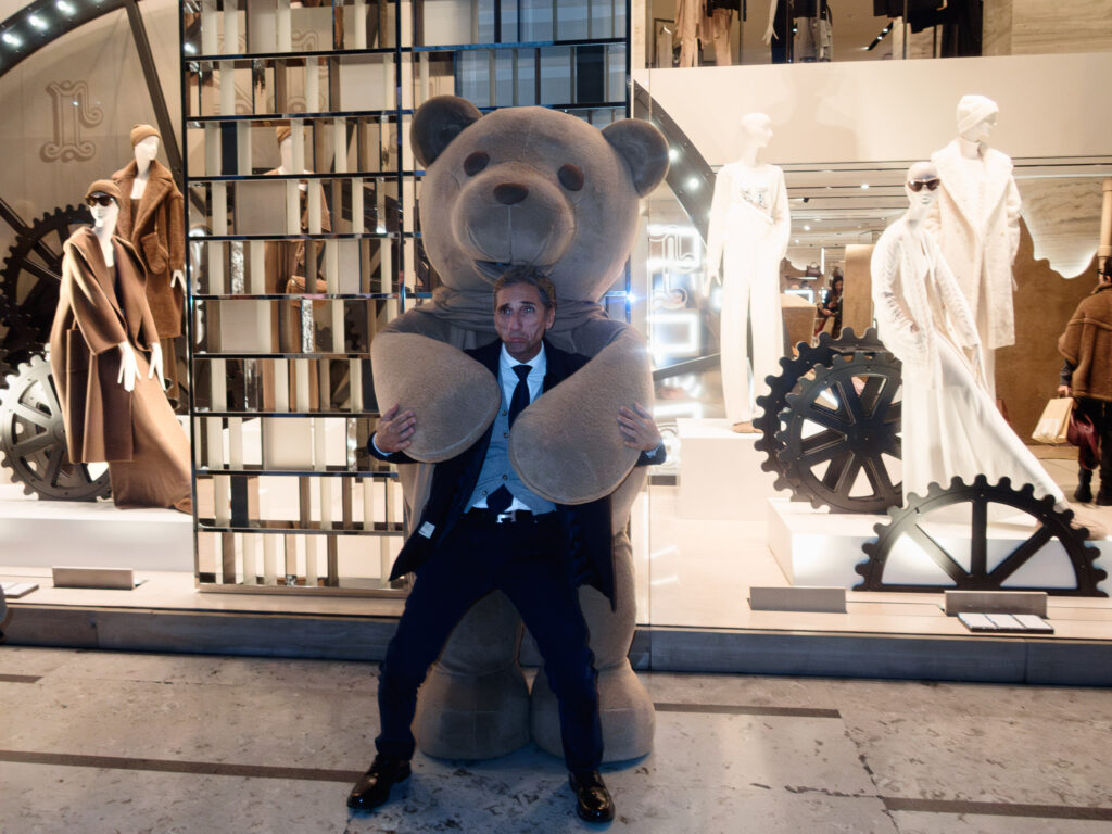 A man in a formal suit poses humorously in front of a large teddy bear statue, appearing to hold its oversized arms. The setting is a high-end fashion store window display featuring mannequins dressed in luxurious clothing, including tailored coats and elegant outfits. The display incorporates mechanical gears and reflective surfaces, adding a modern and industrial aesthetic to the backdrop.