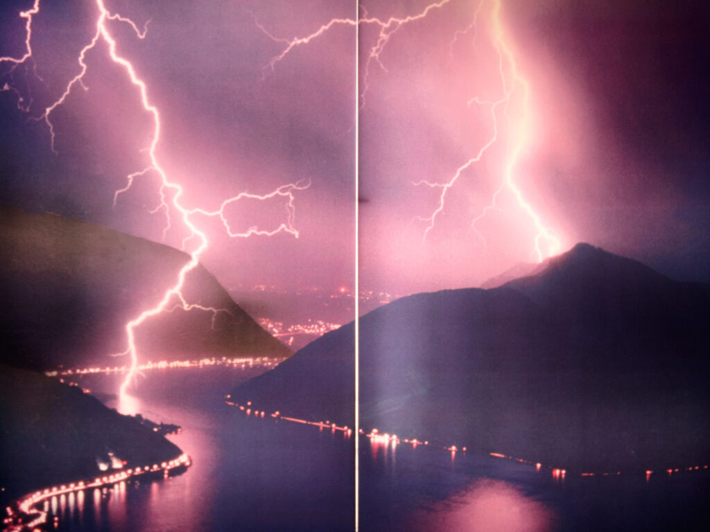 A dramatic nighttime image of two powerful lightning bolts striking over a mountainous landscape, captured on a large printed poster. The vertical line in the center reveals the fold or seam of the poster, indicating it as a physical print. The scene is filled with vibrant purple and pink hues from the lightning, which reflect off the calm waters of a lake below. The shoreline is dotted with glowing lights from towns and roads, blending the raw power of nature with the human presence in the serene landscape.