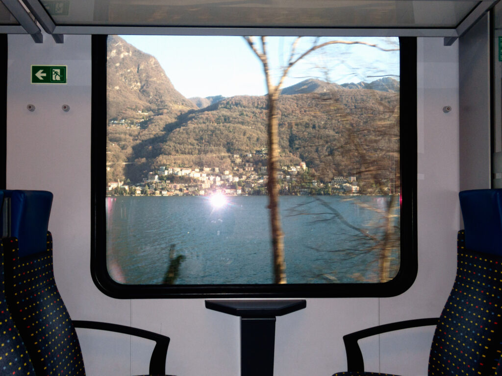 A scenic view of a lake and hillside town framed through the window of a moving train. The landscape features calm blue waters, dense wooded hills, and clusters of buildings along the shoreline under clear skies. A tree trunk appears blurred in the foreground, emphasizing the motion of the train. Inside the train, two empty seats with dark fabric and yellow dotted patterns flank a small table beneath the window, creating a contrast between the dynamic outside view and the still interior.