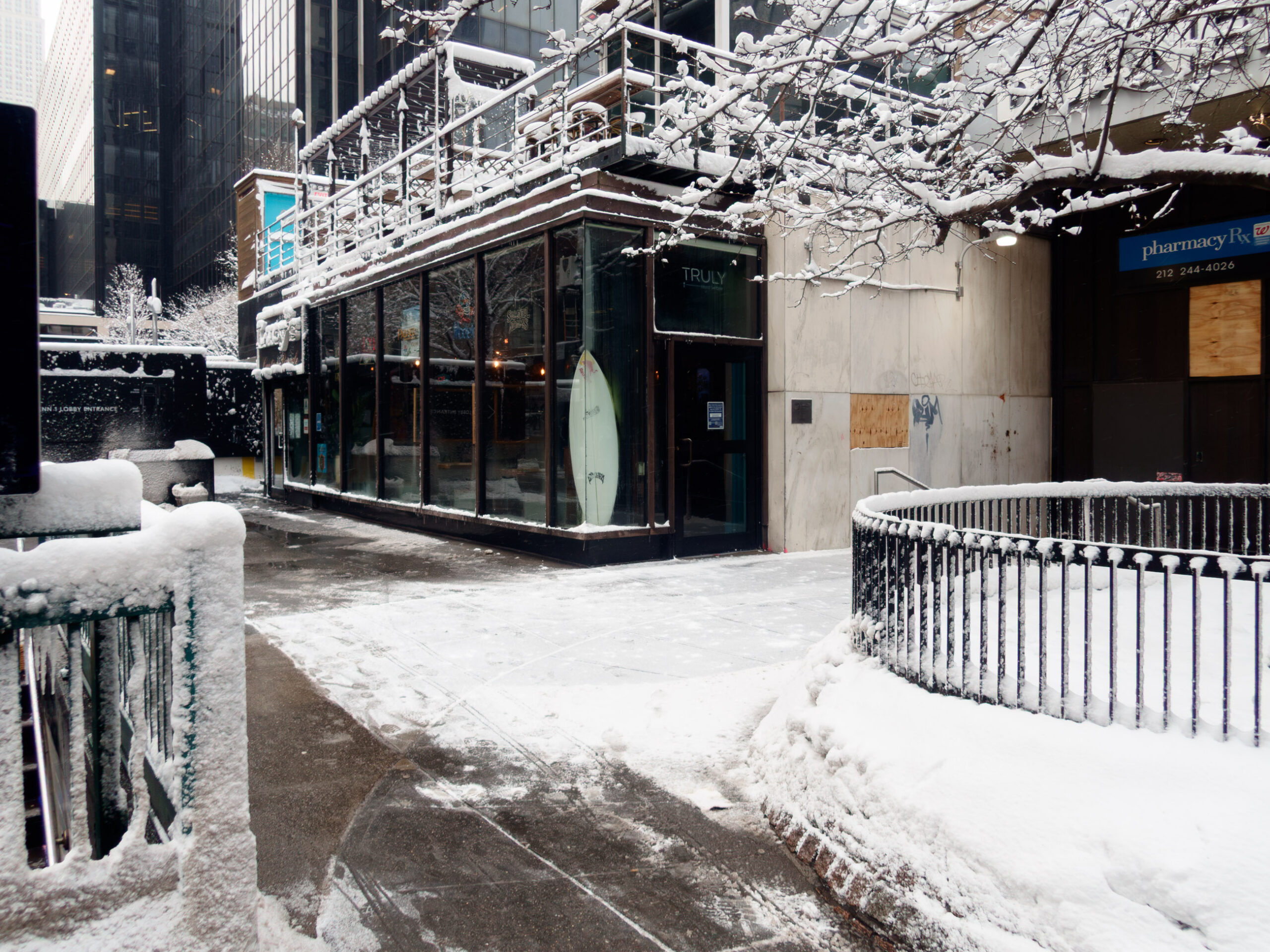 Photograph of a snowy urban scene: A corner restaurant with large glass windows and a surfboard leaning against it is blanketed in snow, with its outdoor terrace covered in a layer of white.