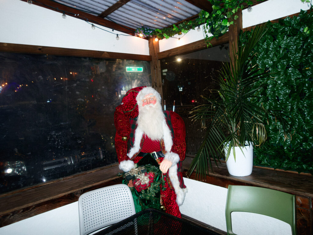 A festive indoor scene featuring a life-sized Santa Claus figure dressed in a traditional red suit with white fur trim, positioned in the corner of a cozy room. The background includes large windows with reflections of nighttime city lights, and the walls are adorned with artificial greenery and string lights. A potted palm adds a tropical touch to the decor, while a simple table and chairs sit in the foreground, completing the inviting, holiday-themed atmosphere.
