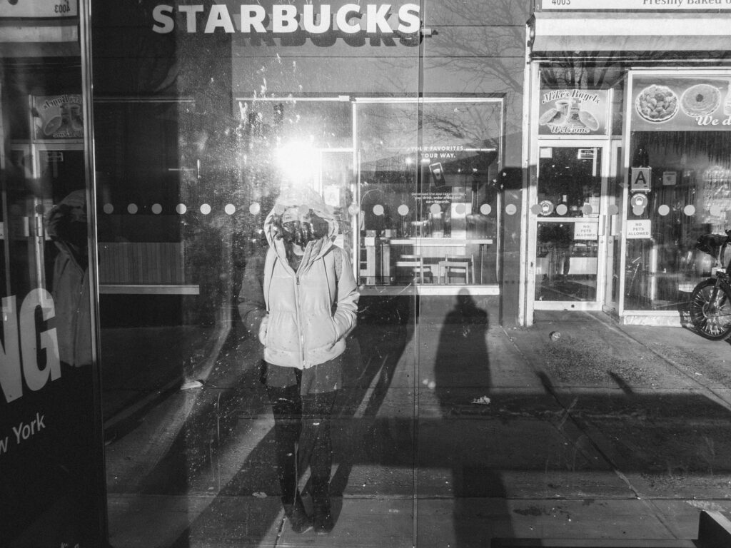 Black and white photograph of a city street scene: A person in a hooded jacket stands in front of a Starbucks storefront, partially reflecting in the glass. Beside it is another storefront advertising freshly baked goods. The sun casts strong shadows on the pavement, highlighting the pedestrian's silhouette and other details on the ground.