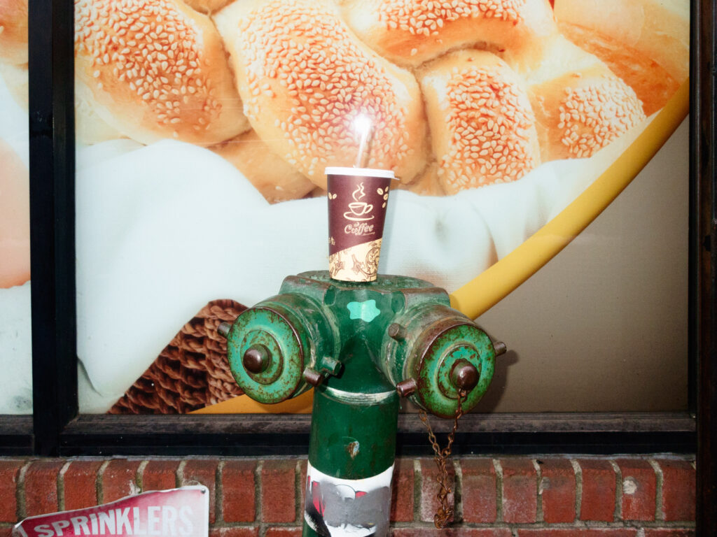 A quirky street photography shot of a green fire hydrant with a disposable coffee cup balanced on top. The backdrop features a large bakery advertisement with close-up images of sesame-covered bread rolls, creating an unexpected contrast between the urban and commercial elements. A brick wall and a partially visible 'SPRINKLERS' sign add texture to the composition, emphasizing the everyday yet artistic details of the scene.